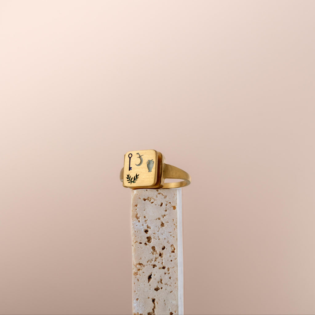 Bague poison chevalière dorée gravée de symboles mystiques présentée sur socle en pierre beige