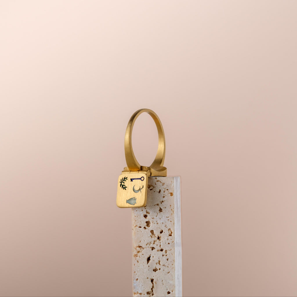 Bague poison dorée avec cadenas gravé de symboles mystiques posée sur socle en terrazzo