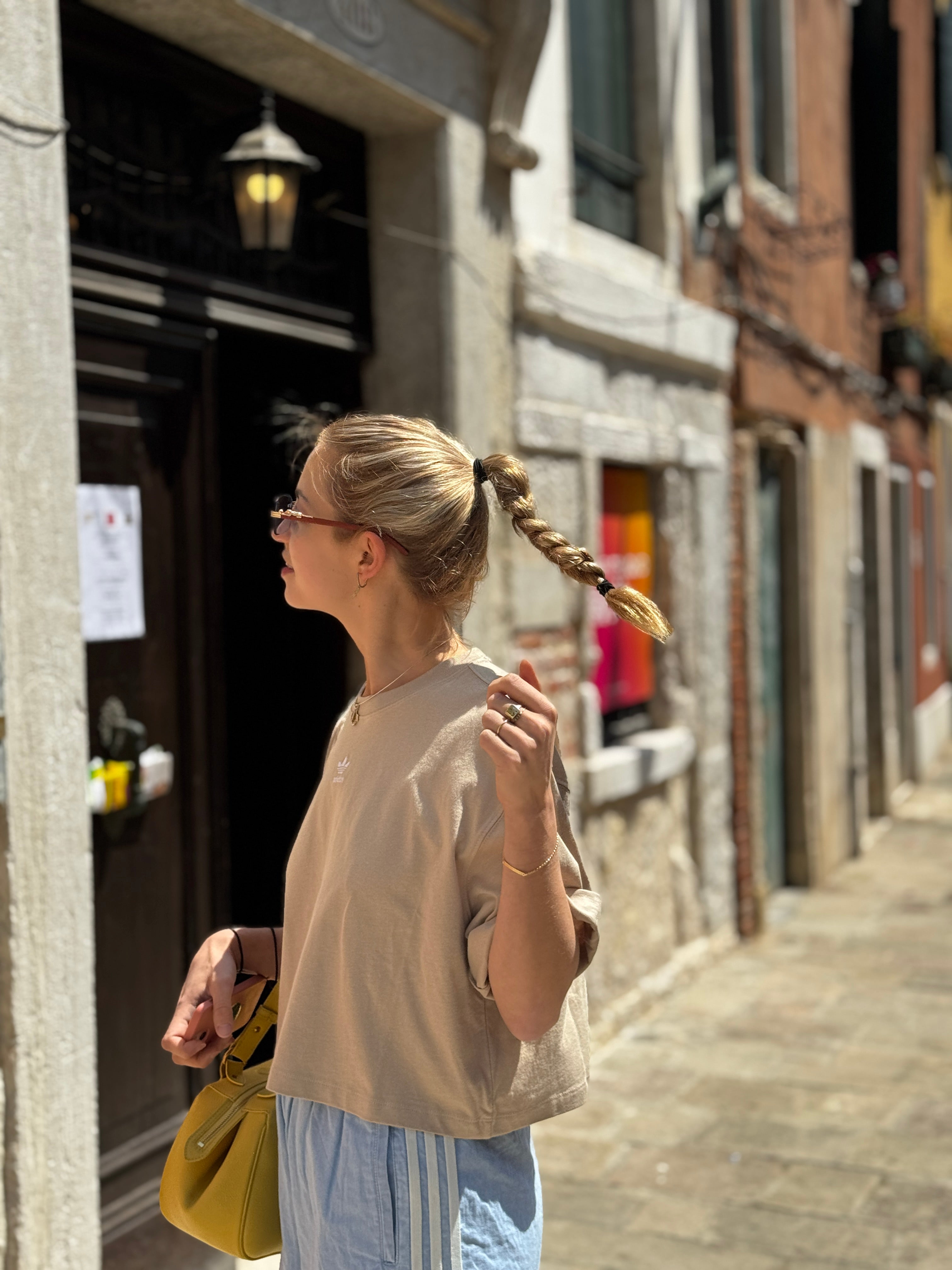 Femme blonde avec tresse longue portant sac jaune et bague poison dans ruelle européenne ensoleillée
